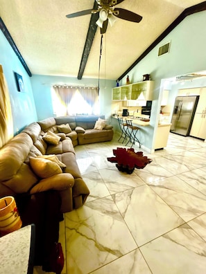 A living room with a sofa, a dining area with chairs, a kitchen with a refrigerator, and a ceiling fan.