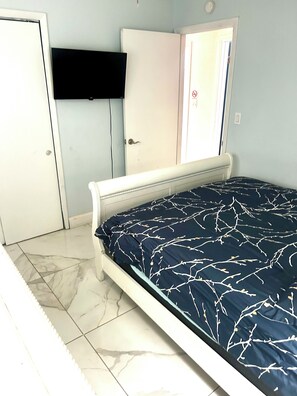 A bedroom with a bed, a door, a wall-mounted TV, and a tiled floor.