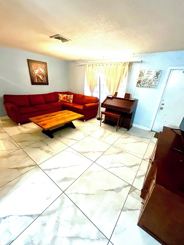 A living room with a red sofa, a wooden coffee table, a piano, and a tiled floor.
