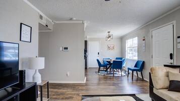 A modern living room with a flat-screen TV, a white lamp, a sofa, and a dining area with blue chairs and a wooden table.
