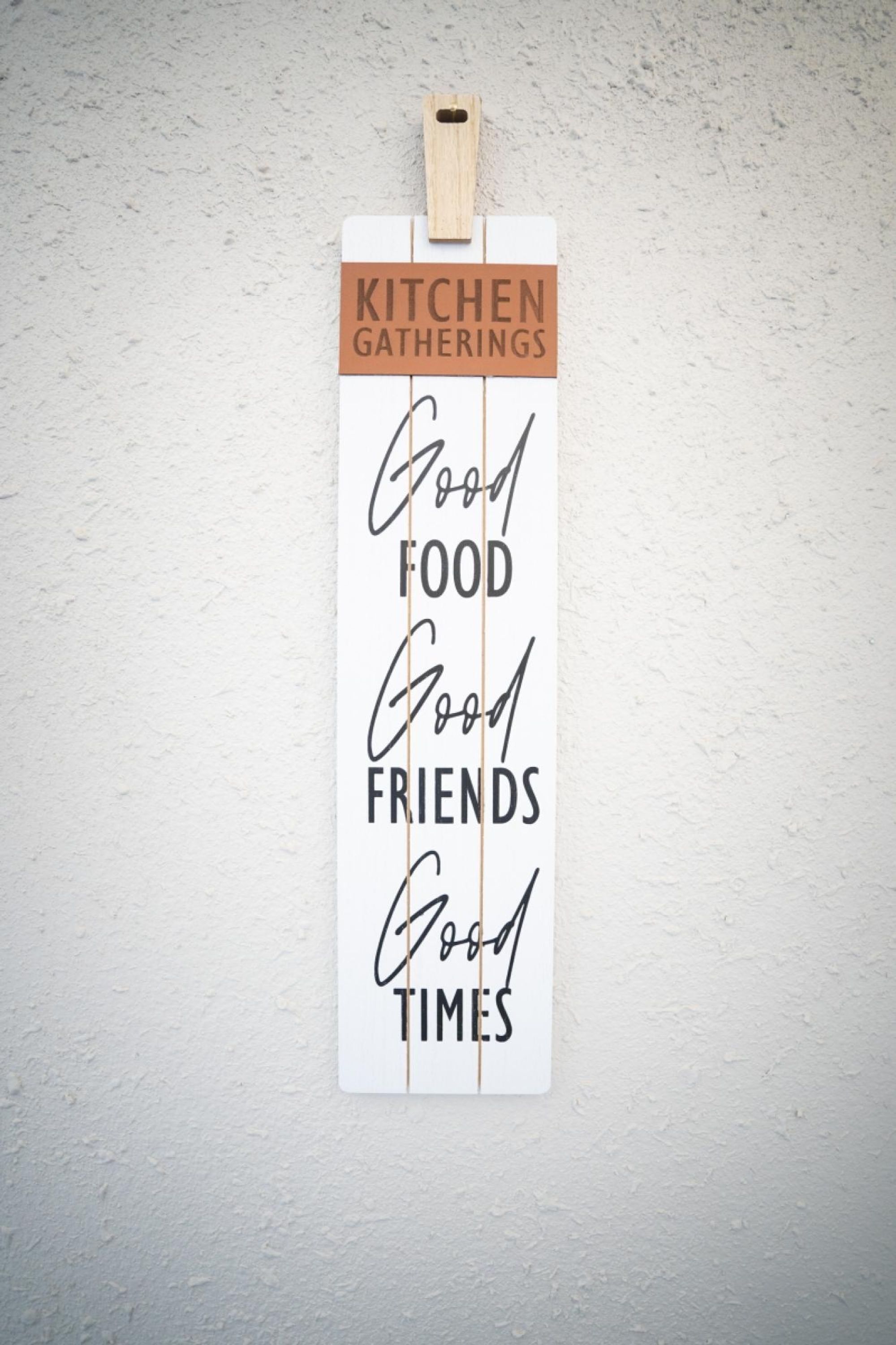 Ein Schild mit dem Text 'Kitchen Gatherings Good Food Good Friends Good Times' vor weißem Hintergrund, oben befestigt mit einer Holzklammer.