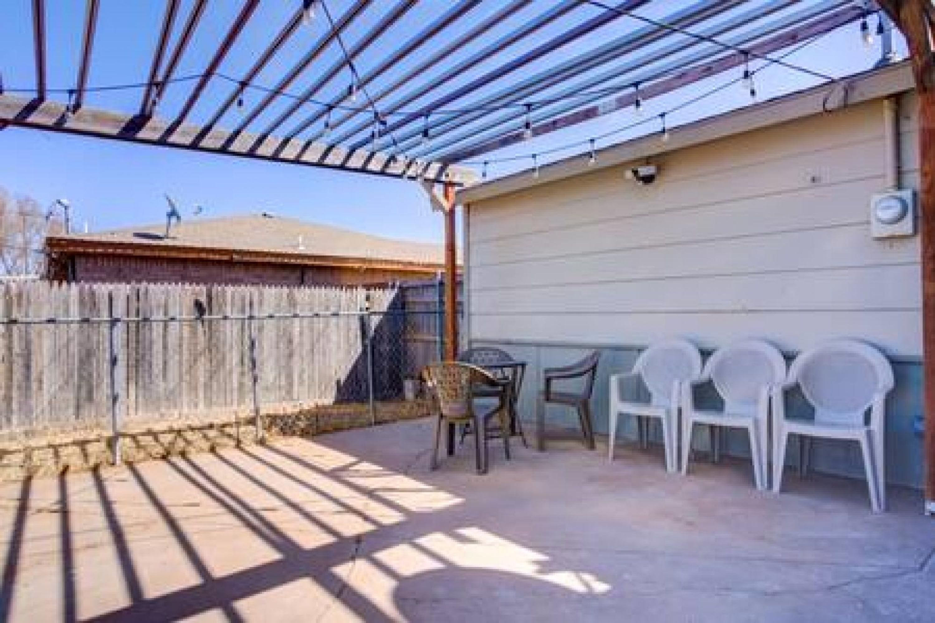 Un patio exterior con pérgola, valla de madera y sillas de plástico.