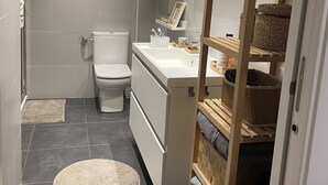 Moderni kylpyhuone, jossa on wc, pesuallas ja suihkutila. Kylpyhuoneessa on hyllyjä, joilla on hygieniatuotteita, ja peili on sijoitettu pesualtaan yläpuolelle.