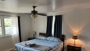 Een slaapkamer met een bed, een plafondventilator, een nachtkastje, een lamp en een raam met gordijnen.