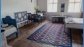 Quarto com cama azul, cadeira azul, mesa com um vaso de flores, tapete estampado e janela com cortinas.