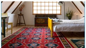 A room with a sloped ceiling, a large window, a bed, a chair, a small table, and a colorful rug. - Viscri 9 (Bunesti)