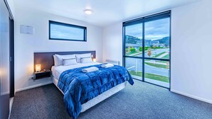 A modern hotel room with a large bed, a nightstand, a bedside lamp, and a window offering a view of the outside. - Amazing Alpine Views in Wanaka (Wānaka)