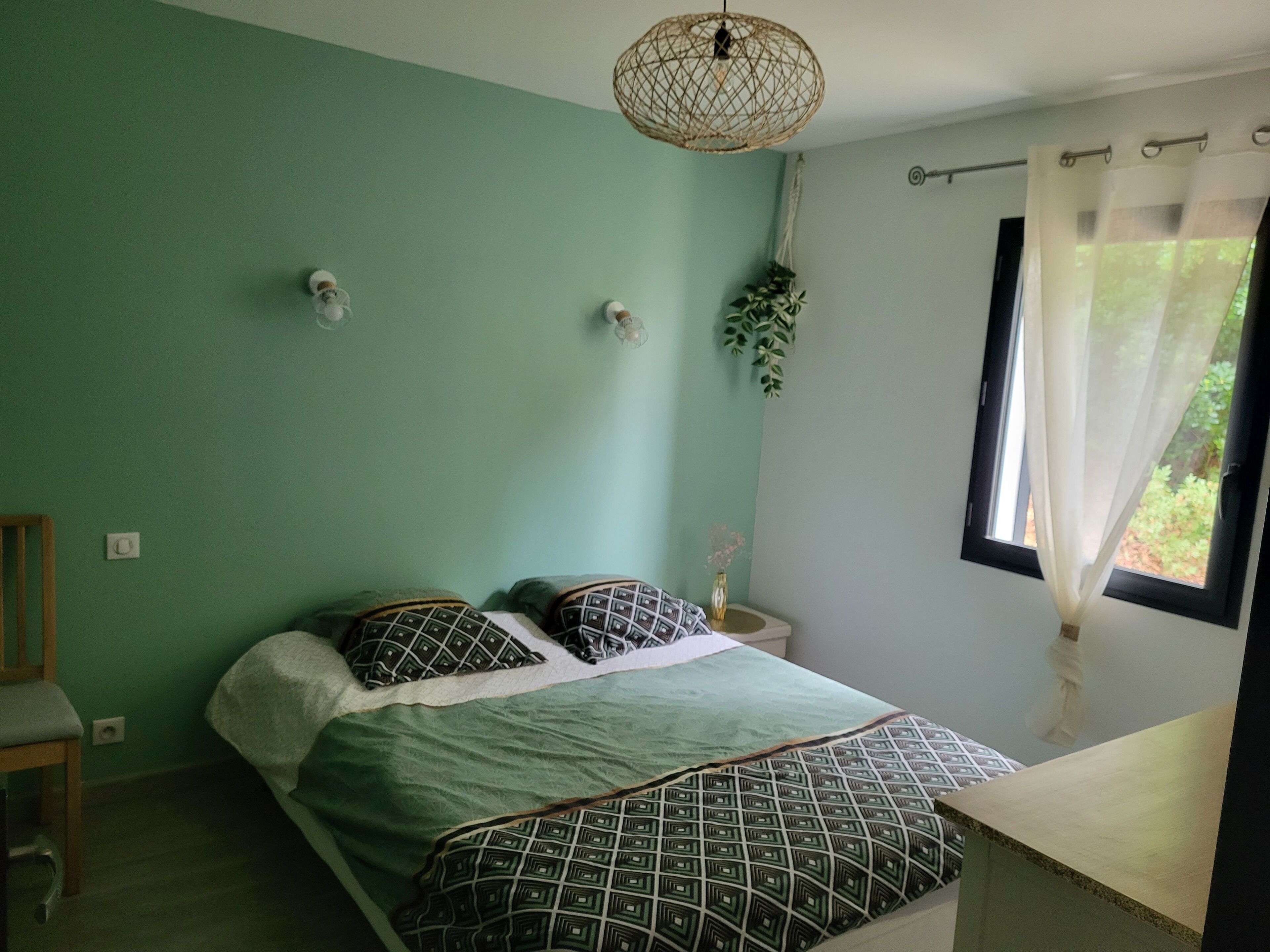 Une chambre à coucher avec un lit, une table de chevet, une plante suspendue et une fenêtre avec des rideaux.