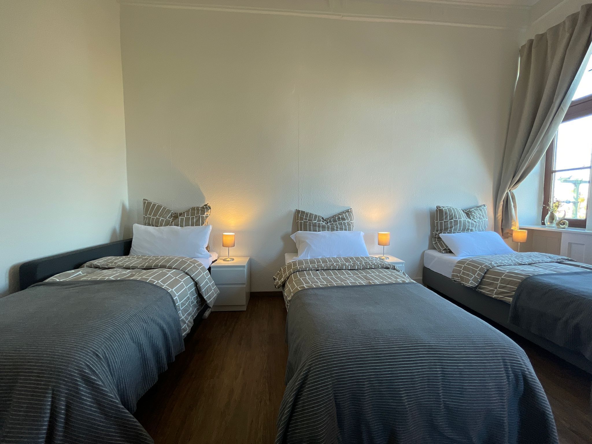A hotel room with two single beds, each with a nightstand and a lamp. The room has a wooden floor and white walls.