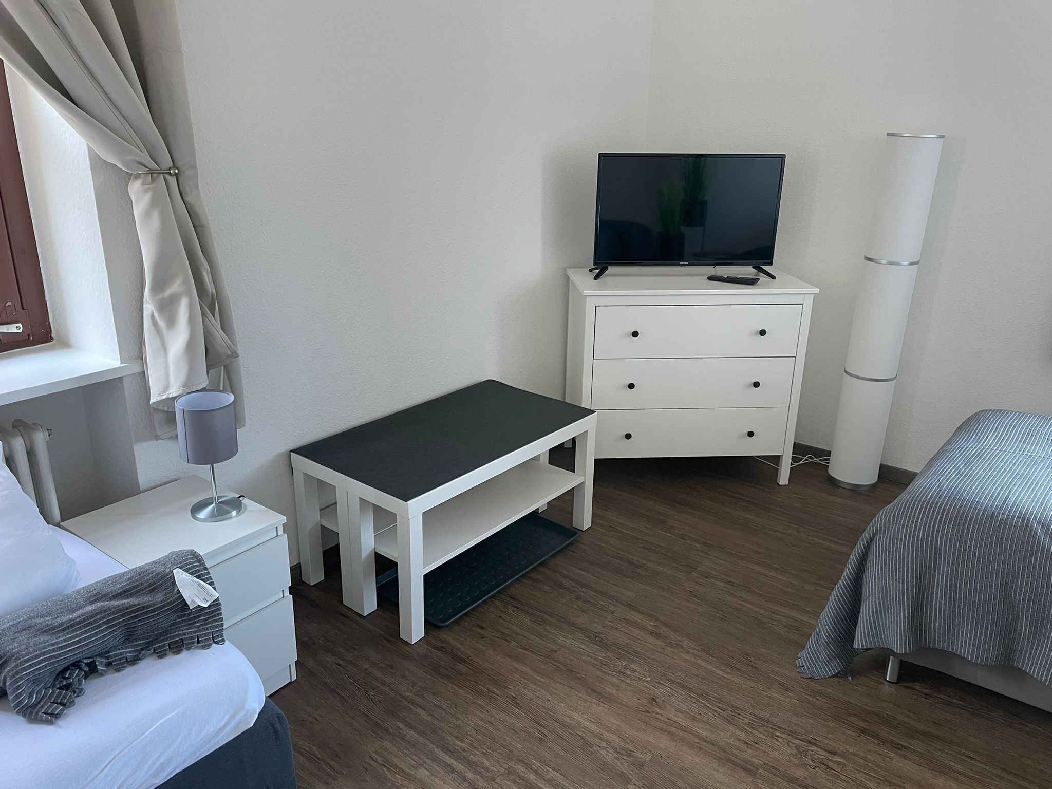 Et hotelværelse med en seng, et lille skrivebord med lampe, et fjernsyn på en kommode og et sengebord.