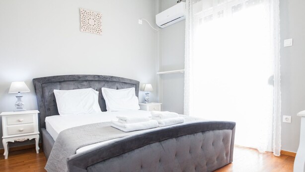 A neatly arranged bedroom with a grey upholstered bed, white bedding, two bedside tables with lamps, and an air conditioning unit mounted on the wall. - Modern apt with Increbilbe Athens Views (Galatsi)