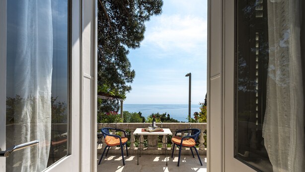 Een open deur die leidt naar een balkon met twee stoelen en een tafel, met uitzicht op een prachtig zeegezicht.