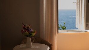 Een kamer met een ronde tafel, een stoel, een vaas met bloemen, een ingelijst kunstwerk aan de muur en uitzicht op zee door een open raam.