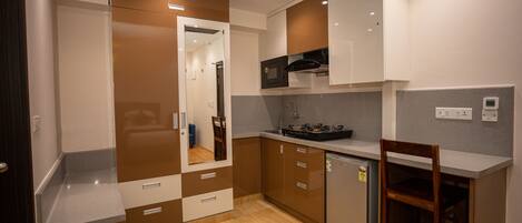 Una cocina compacta con microondas, estufa y horno empotrado. Hay un refrigerador pequeño y una silla de madera. La cocina tiene piso de madera y armarios blancos.