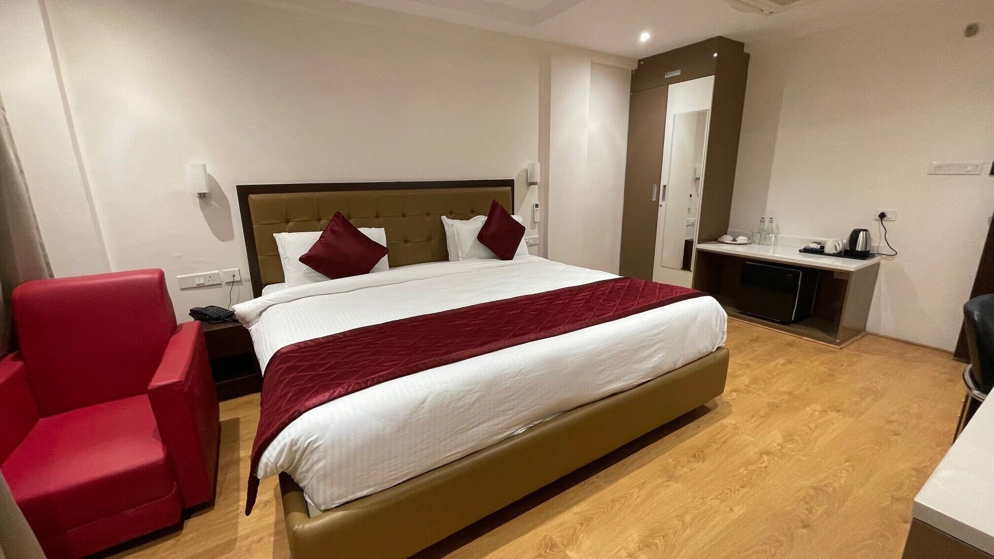 A hotel room with a bed, a red chair, a desk with a mirror, and a small kitchenette.