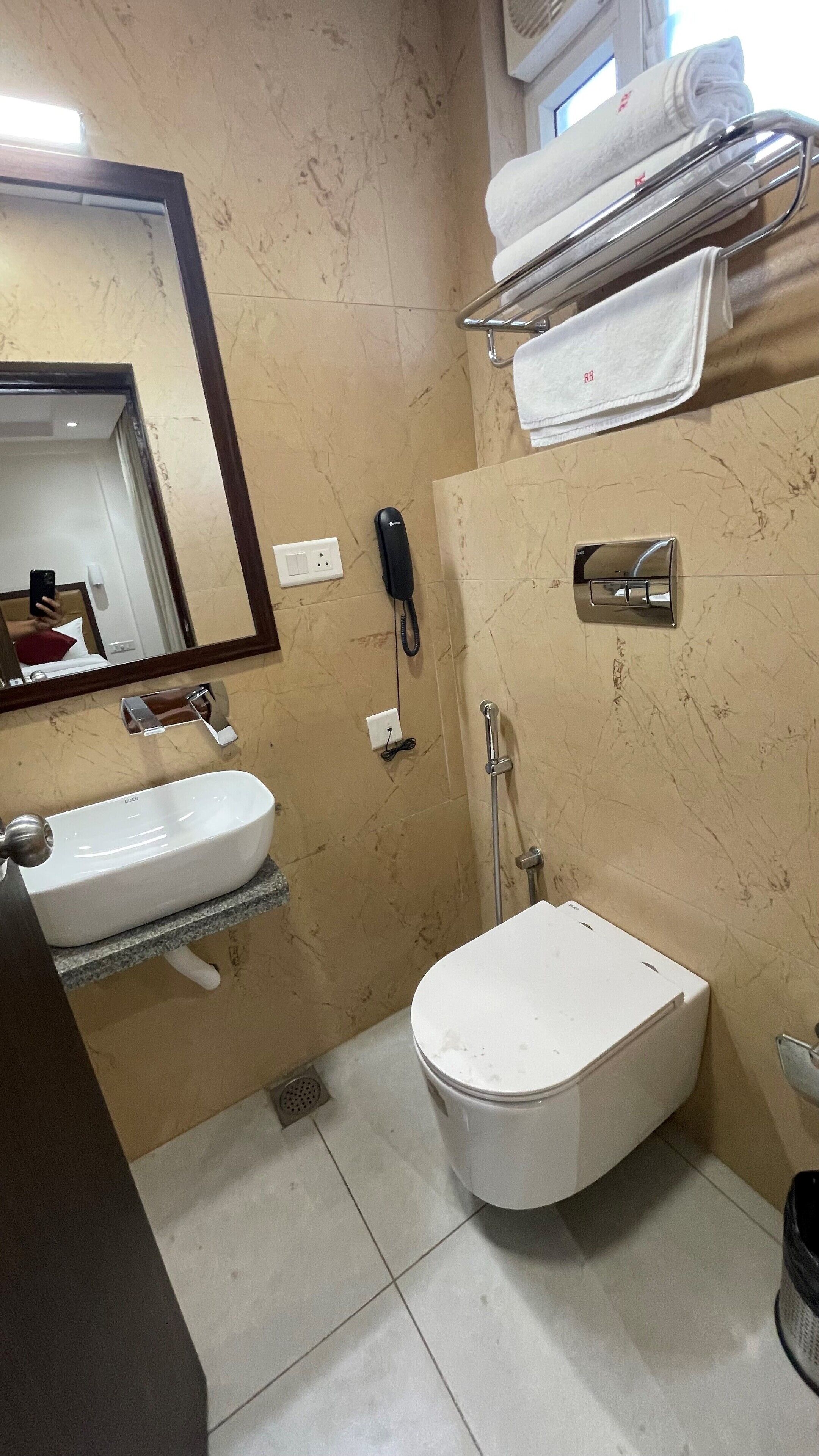 A bathroom with a sink, toilet, and shower. There is a mirror, a towel rack, and a phone on the wall.