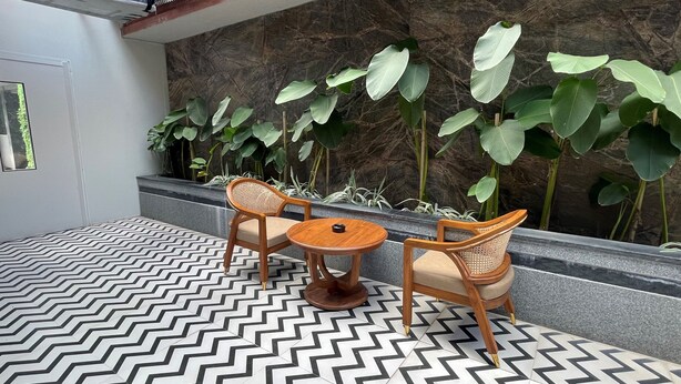 A seating area with two wooden chairs and a small round table, surrounded by large green plants and a patterned floor. - Hotel Silver Clé Hitech City (Hyderabad)