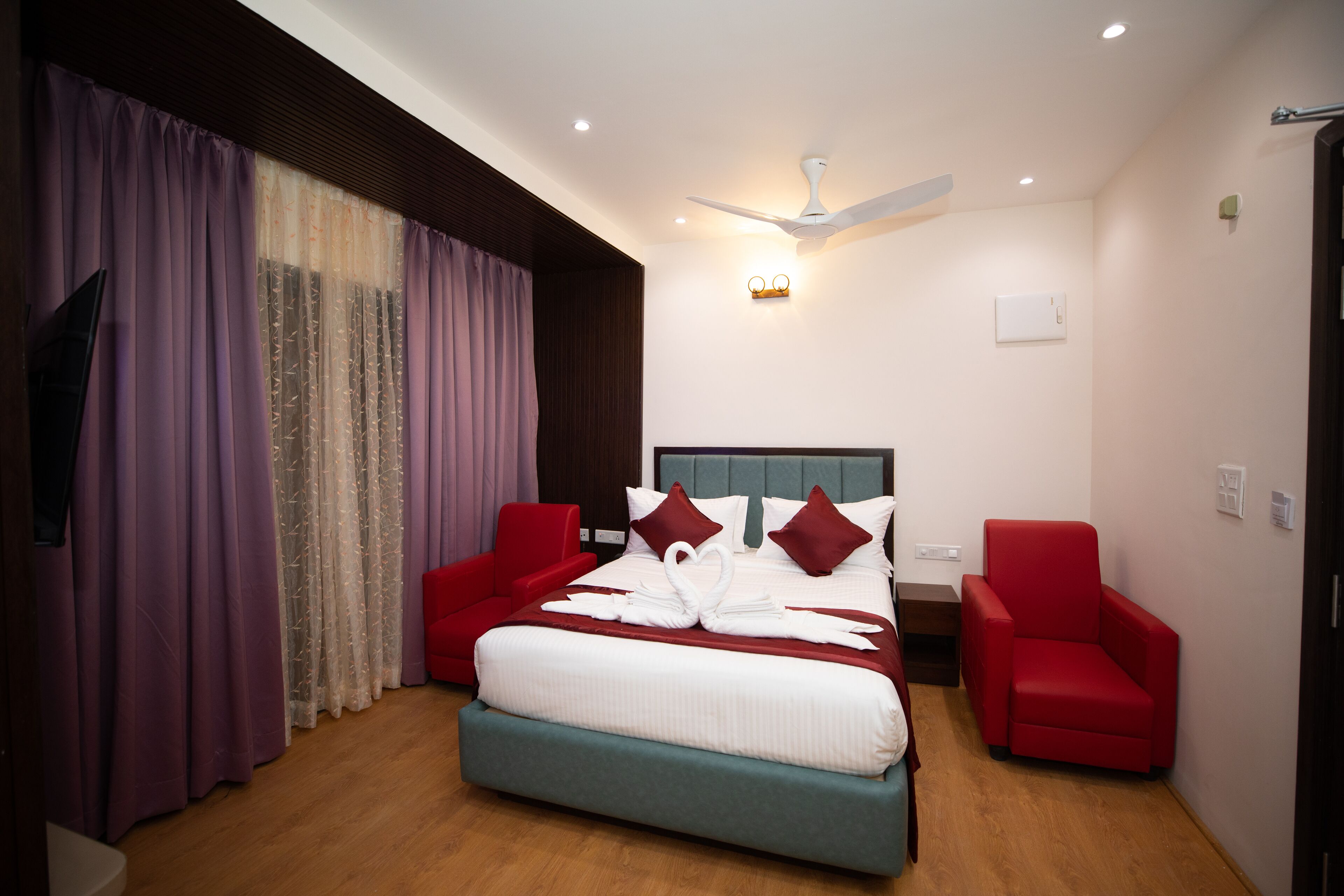 Une chambre d’hôtel moderne avec un grand lit, deux fauteuils rouges, un ventilateur de plafond et une télévision.