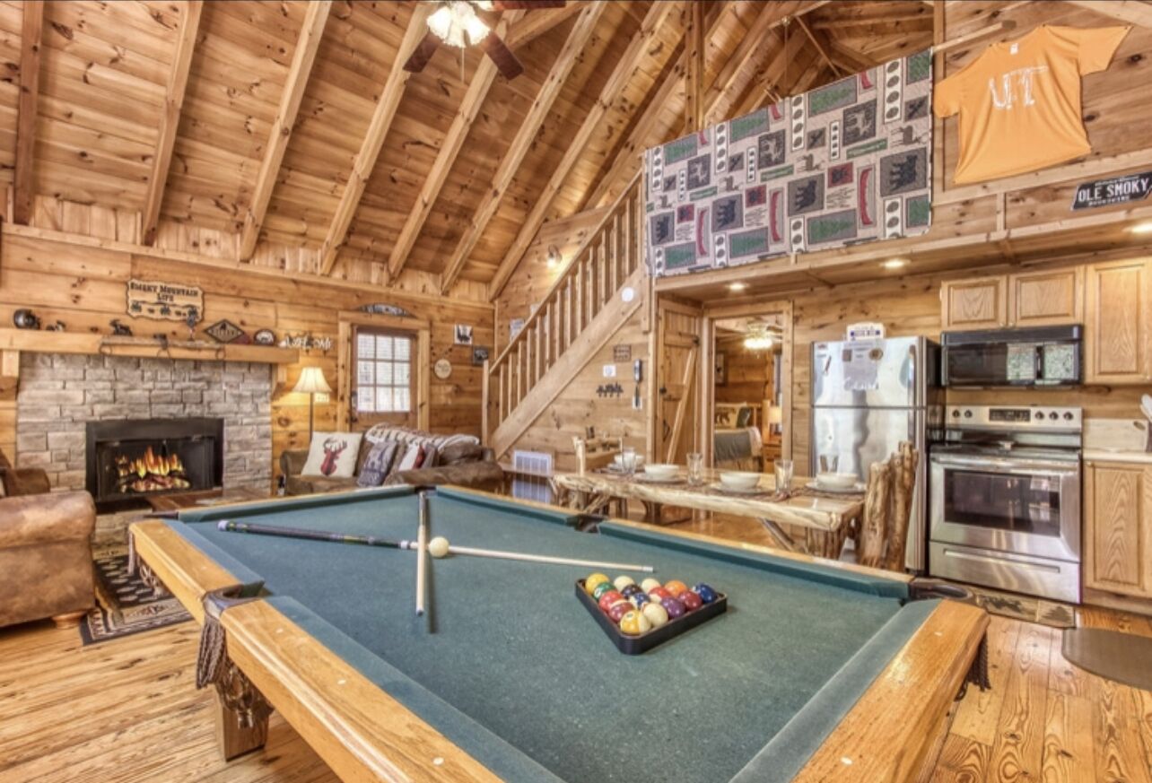 A wooden cabin interior with a pool table, fireplace, kitchen, and a staircase.