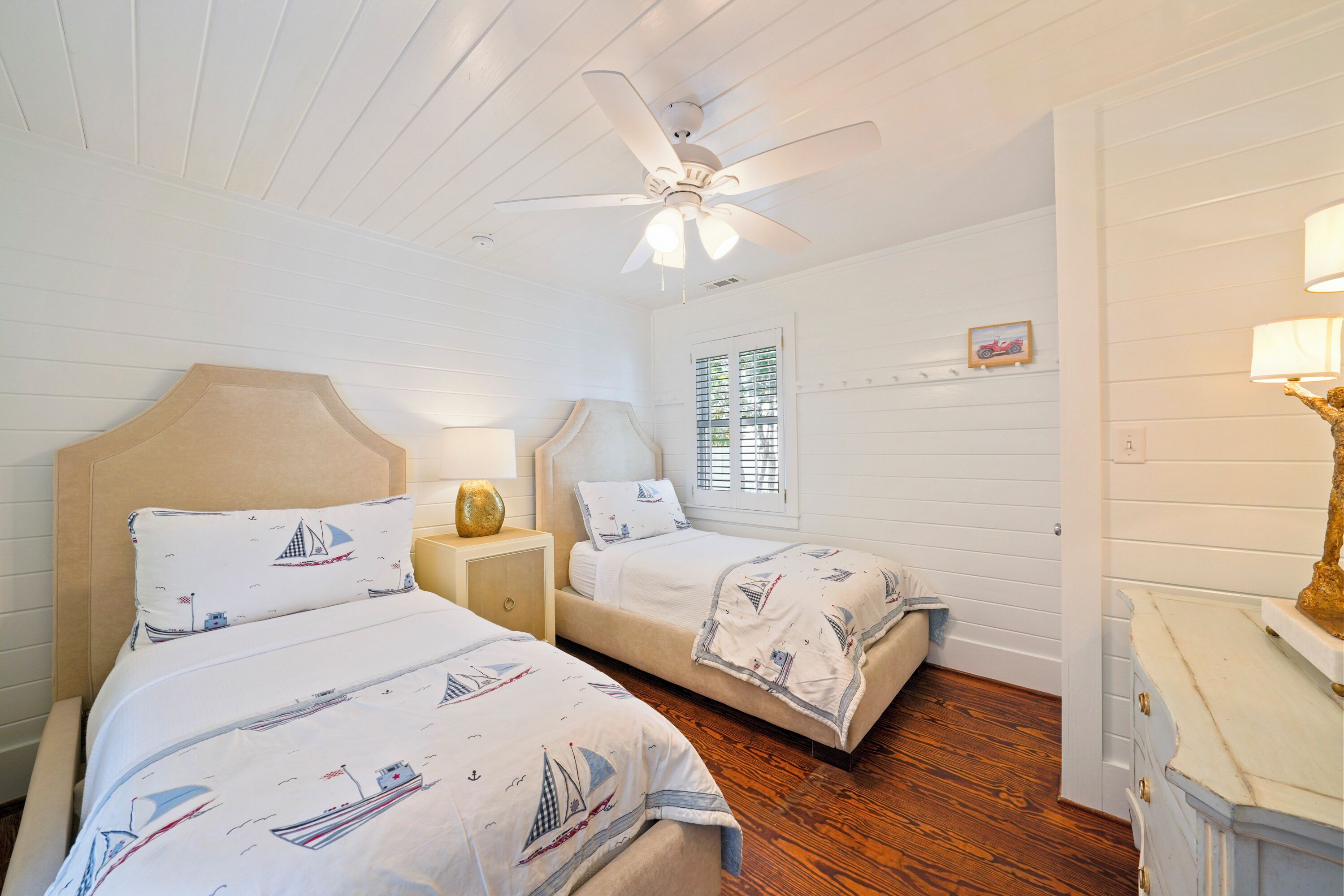 A bedroom with two beds, a ceiling fan, a window with blinds, and a dresser.