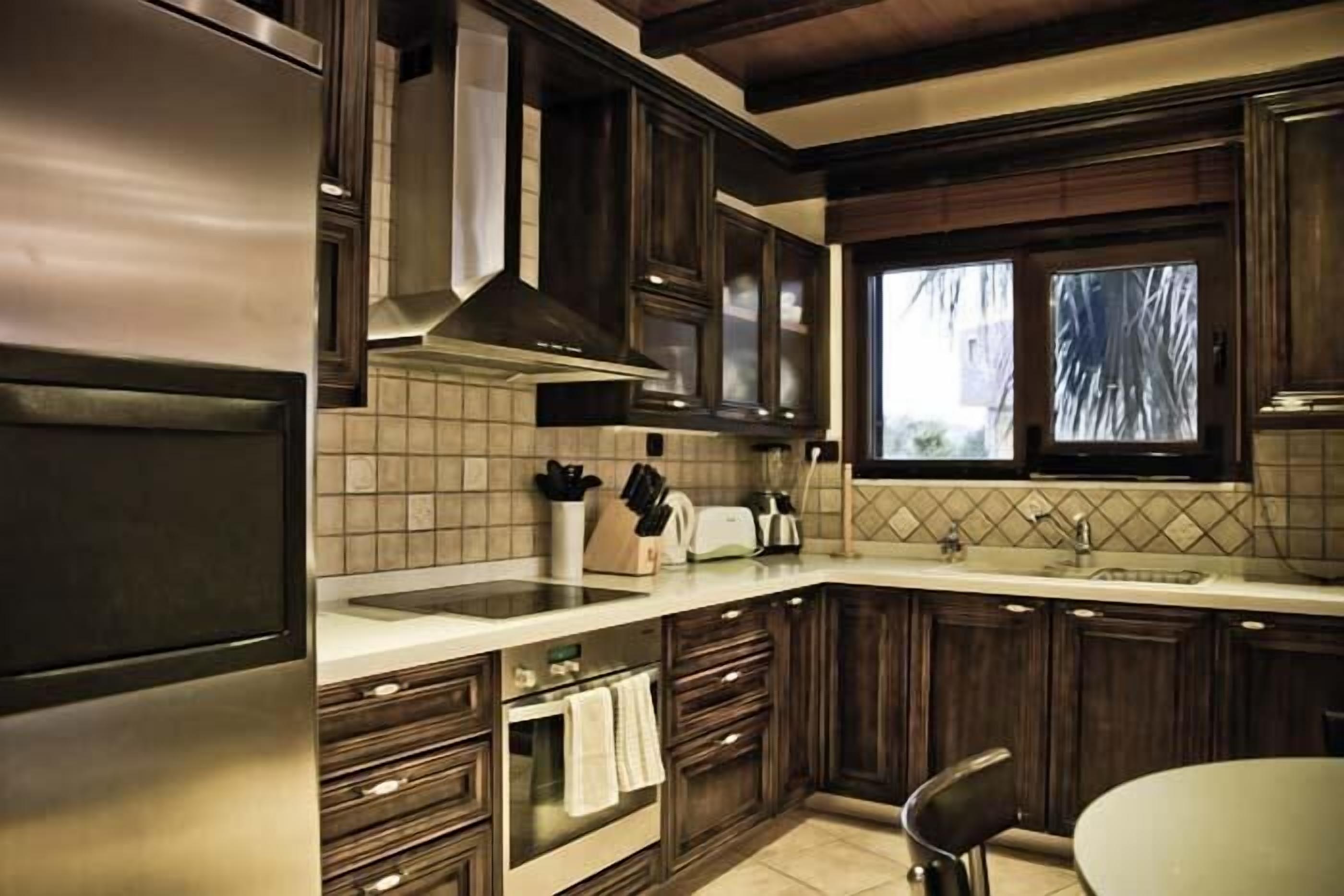 A modern kitchen with dark wooden cabinets, a stainless steel refrigerator, and a window with a view of trees.