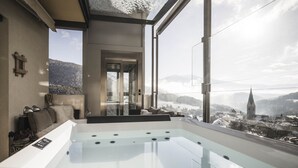 A modern spa area with a glass-enclosed hot tub, a cozy seating area with cushions, and a scenic view of a snowy landscape and a church spire. - Dolomites Alpin Penthouse 90 qm with privat sauna and privat whirlpool, WLAN (Rio di Pusteria)