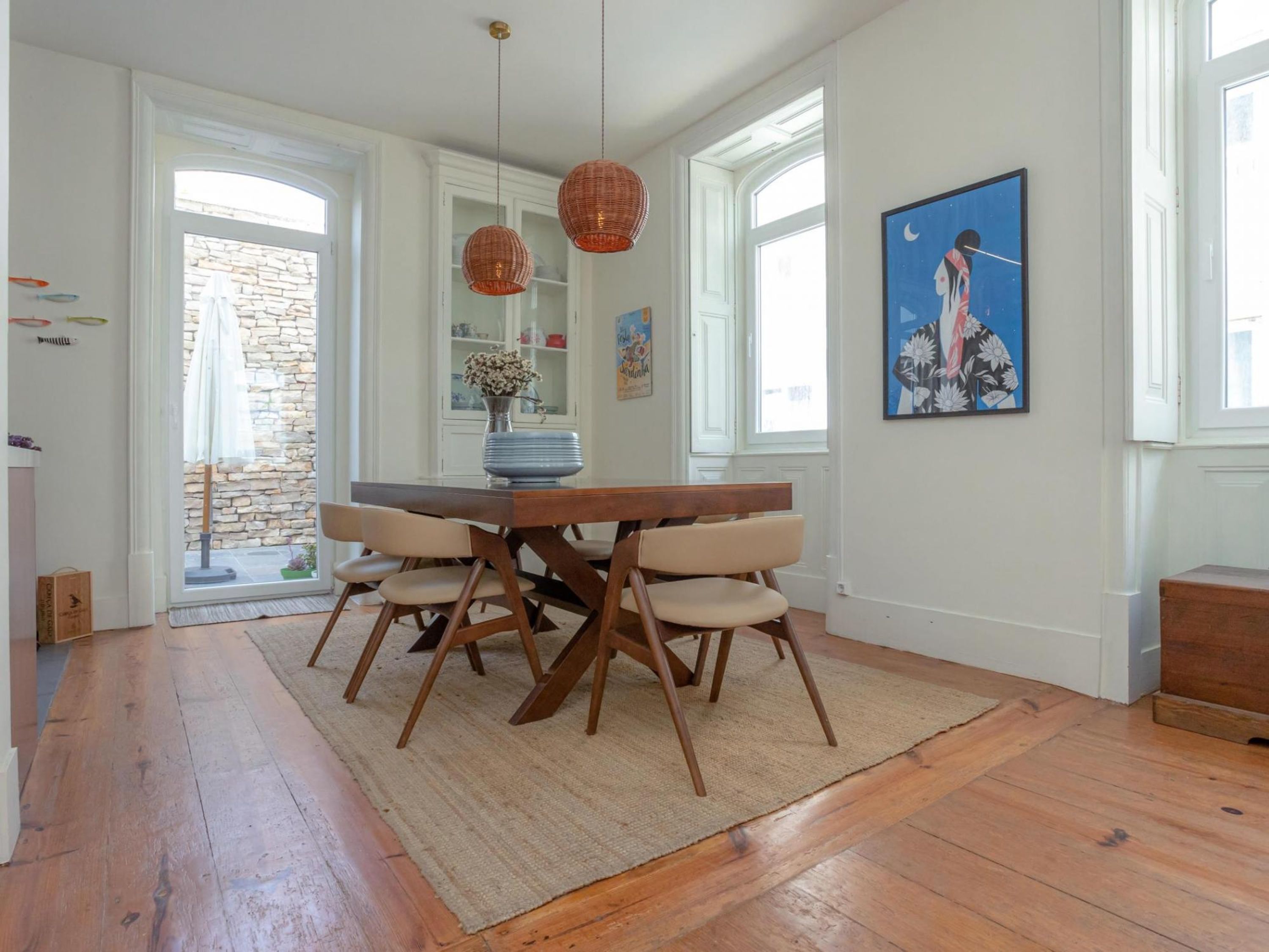 A dining area with a wooden table and chairs, a woven pendant light, and a painting on the wall.
