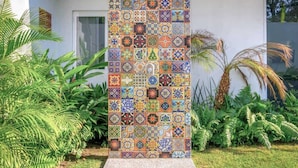 Un mur en mosaïque verticale, composé de différents motifs de carreaux, entouré d’une végétation luxuriante et reposant sur une base en béton.