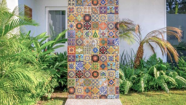 Pared vertical de mosaicos con diversos diseños de baldosas, rodeada de abundante follaje y una base de concreto.