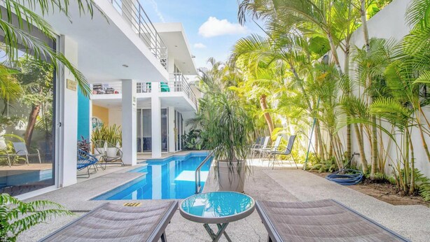 Un área de piscina moderna con sillones de descanso, una piscina y plantas tropicales.