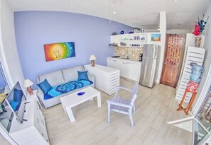 Una sala de estar moderna con un sofá, una mesa de centro y un televisor, una cocina con armarios blancos y un comedor con una silla.