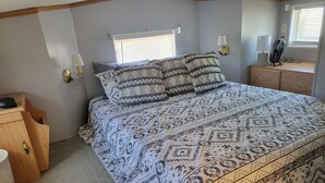 A bedroom with a patterned bedspread, a wooden dresser, a nightstand, and a window with blinds.