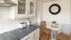 A kitchen with white cabinets, a glass-fronted cabinet, a toaster, a clock, and a small table with chairs. - Cosy Seaside Home with Free Parking - Pass the Keys (North Shields)