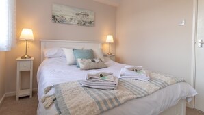 A neatly made bed with pillows and a folded towel, two bedside lamps, a painting on the wall, and a door to the right. - Cosy Seaside Home with Free Parking - Pass the Keys (North Shields)
