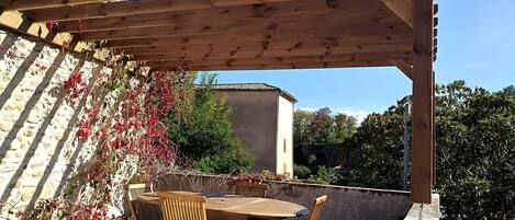 A wooden pergola with a table and chairs underneath, surrounded by greenery and a stone wall.
