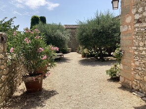 A gravel pathway leading to a stone building with a tiled roof. There are potted plants and a bench along the path.