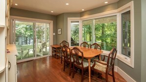 A dining area with a wooden table and chairs, large windows with a view of greenery, and a ceiling with recessed lighting. - Panorama Cottage - perfect sunsets! (Parry Sound)
