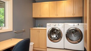 A laundry room with two front-loading washing machines, wooden cabinets, a sink, and a window. - Panorama Cottage - perfect sunsets! (Parry Sound)