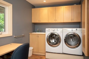A laundry room with two front-loading washing machines, wooden cabinets, a sink, and a window. - Panorama Cottage - perfect sunsets! (Parry Sound)