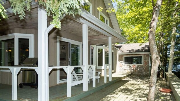 A multi-level wooden deck with a covered area, outdoor seating, and a stone building in the background. - Panorama Cottage - perfect sunsets! (Parry Sound)