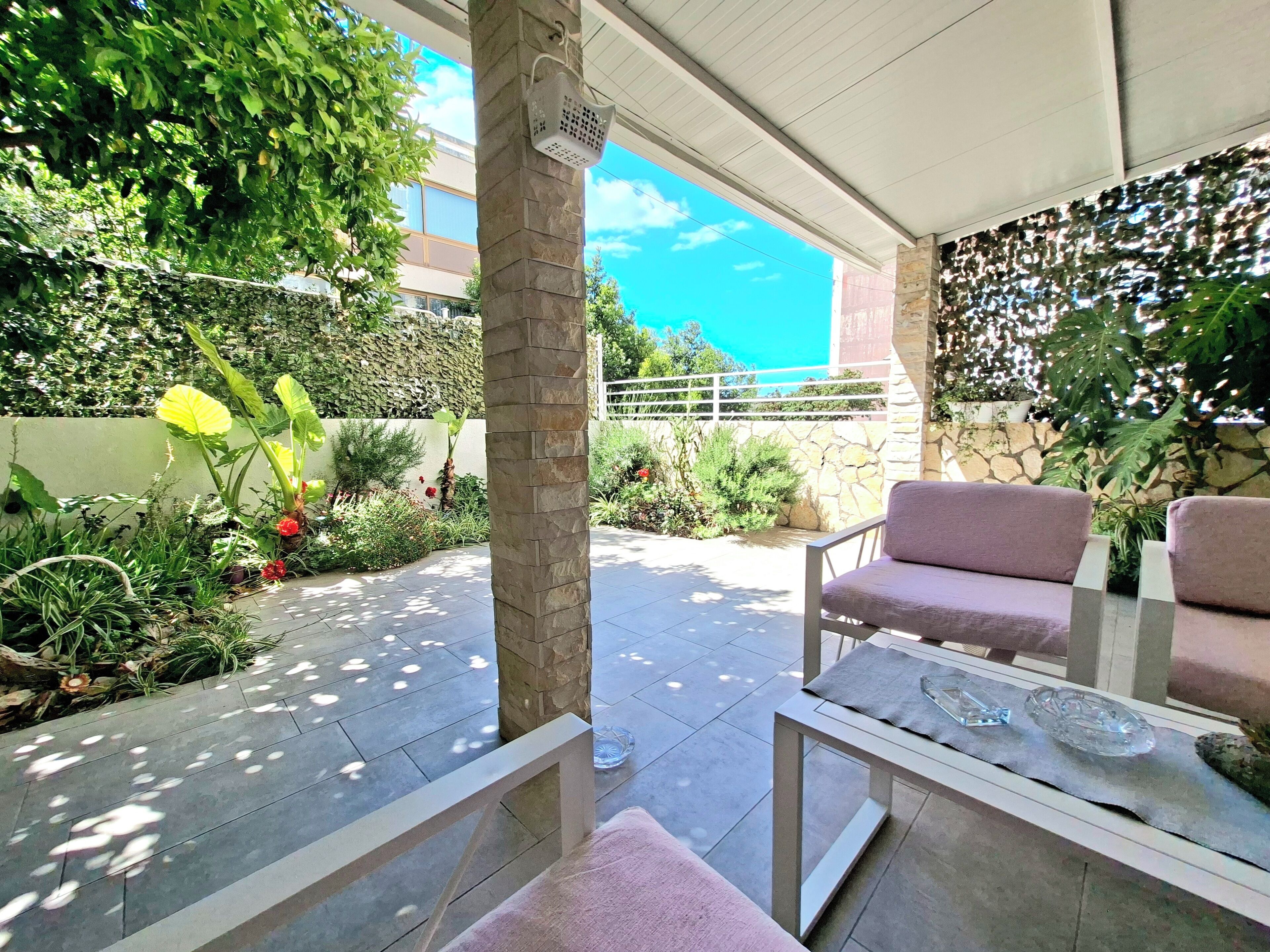A covered outdoor seating area with stone pillars, a table with a glass top, and a variety of plants and flowers.