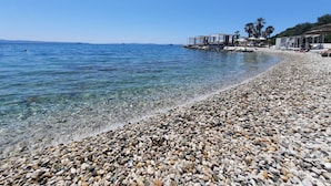Ein Kiesstrand mit klarem Wasser, ein Steg mit Gebäuden und Palmen.