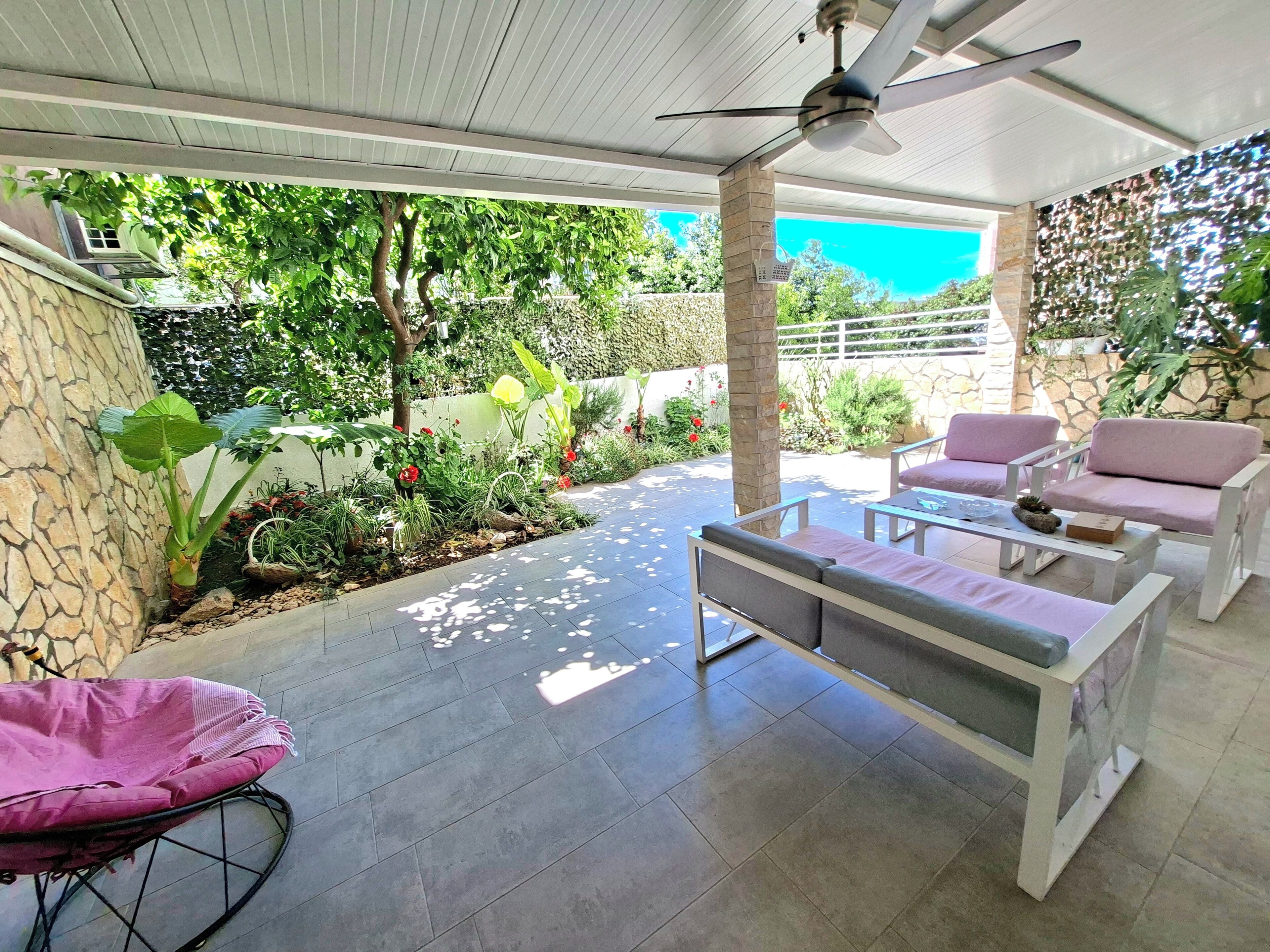 A covered patio area with a ceiling fan, outdoor seating, and a garden with various plants.