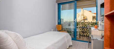 A hotel room with a bed, a chair, a table, and a balcony with a view of buildings and greenery.