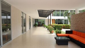 Een moderne, overdekte gang met glazen deuren, potplanten en een zithoek met oranje kussens.