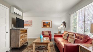 Una sala de estar con un sofá de cuero rojo, una mesa de centro de madera, un televisor sobre un mueble de madera y una ventana con persianas.