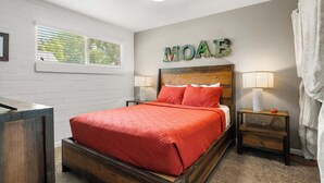 A bedroom with a wooden bed, a nightstand with a lamp, a window with blinds, and a wall-mounted sign.