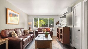 Ein Wohnzimmer mit braunem Ledersofa, einem Holztisch und einem Schrank. Ein Fenster mit Jalousien, eine Lampe und ein gerahmtes Bild sind an der Wand zu sehen.