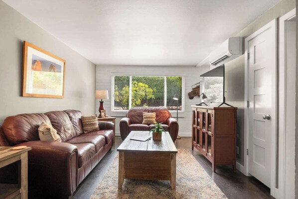 A living room with a brown leather sofa, a wooden coffee table, and a cabinet. There is a window with blinds, a lamp, and a framed picture on the wall.
