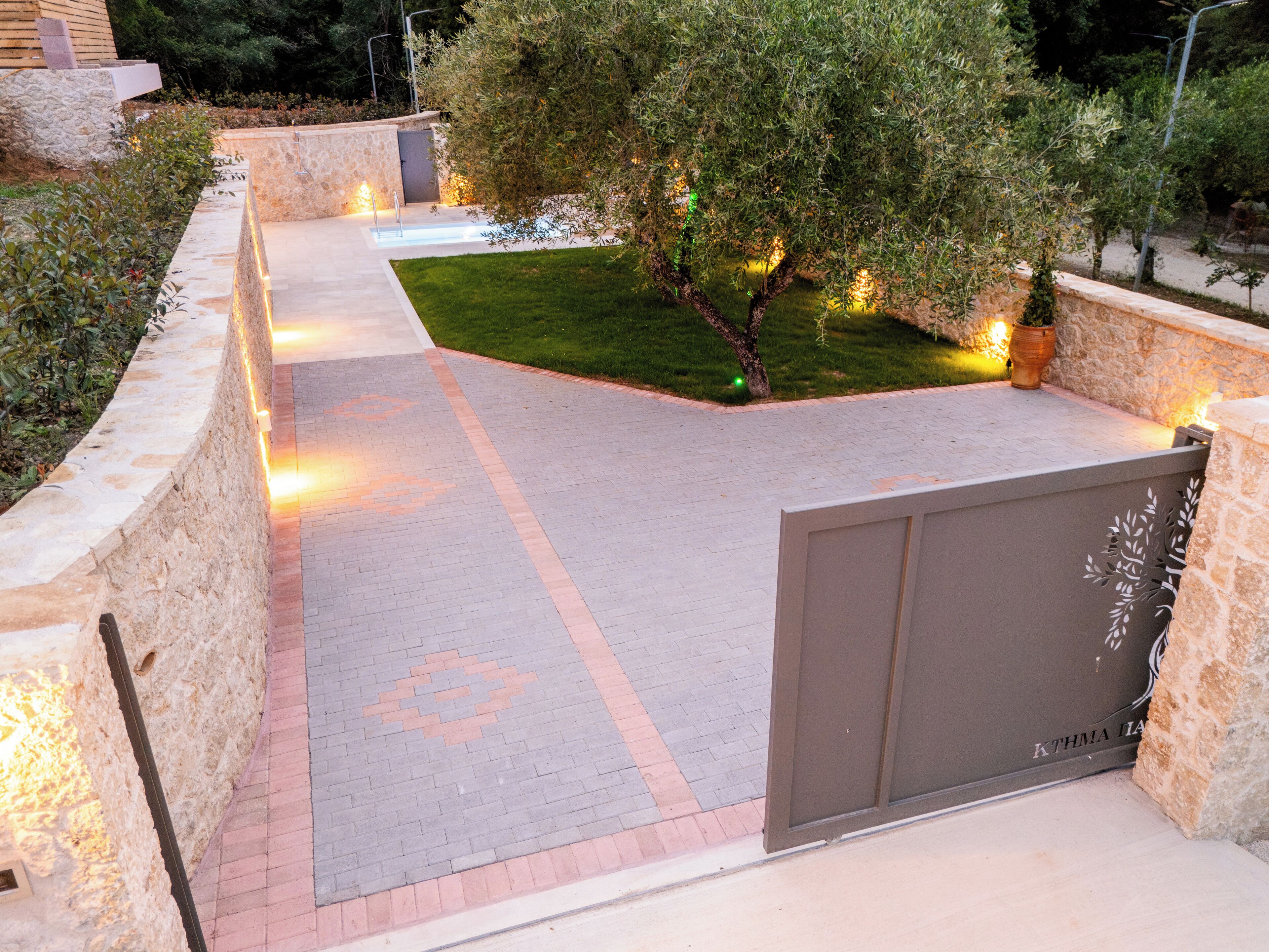 Un chemin pavé mène à une entrée fermée par un portail, avec un mur de pierre d’un côté et une zone herbeuse de l’autre.